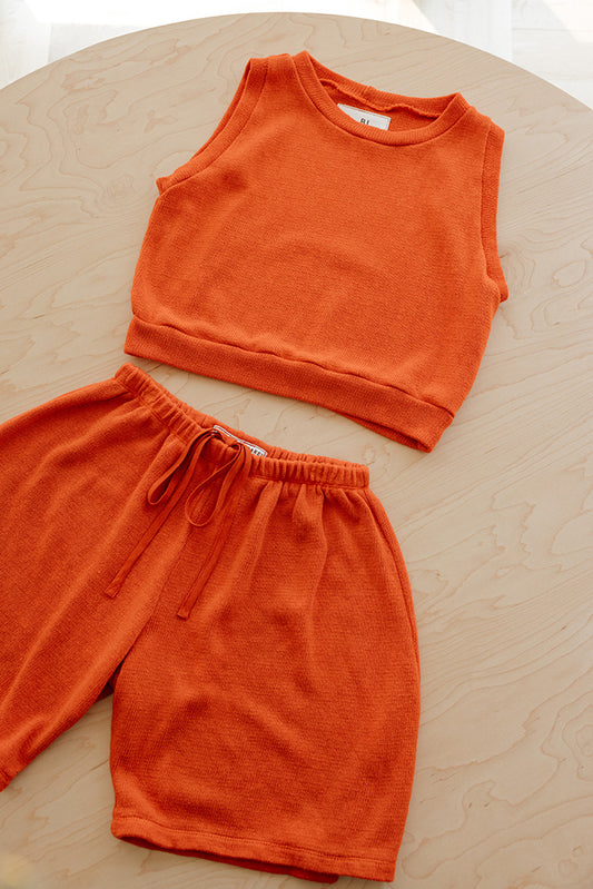 An orange two-piece loungewear set consisting of a sleeveless cropped tank top and matching drawstring shorts, laid flat on a light wood-grained circular table.